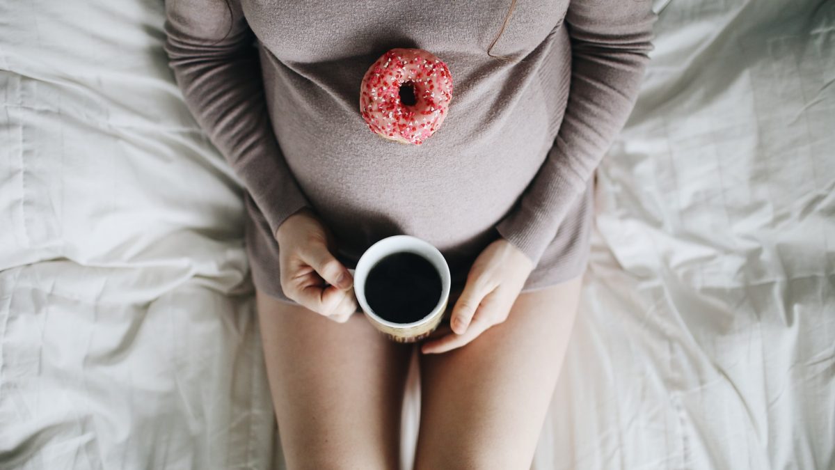 Overhead image of pregnant woman enjoying tea with a donut atop her stomach